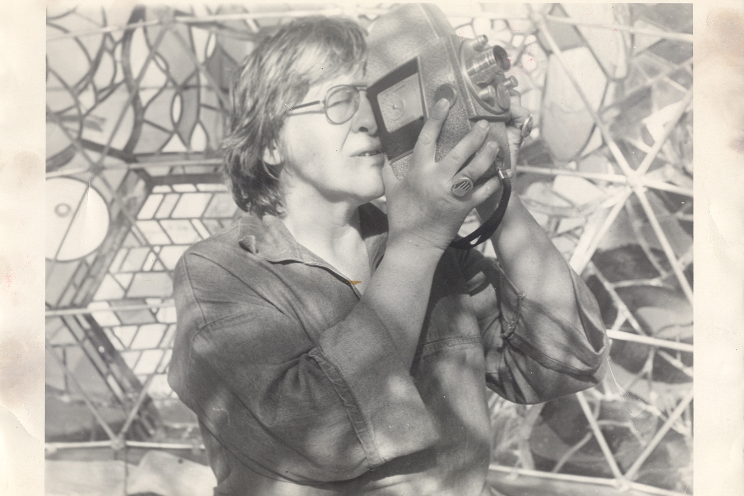 A black and white image of Phyllis Birkby, a woman with short hair and glasses, looking through a film camera. The background is a geometric tile pattern.