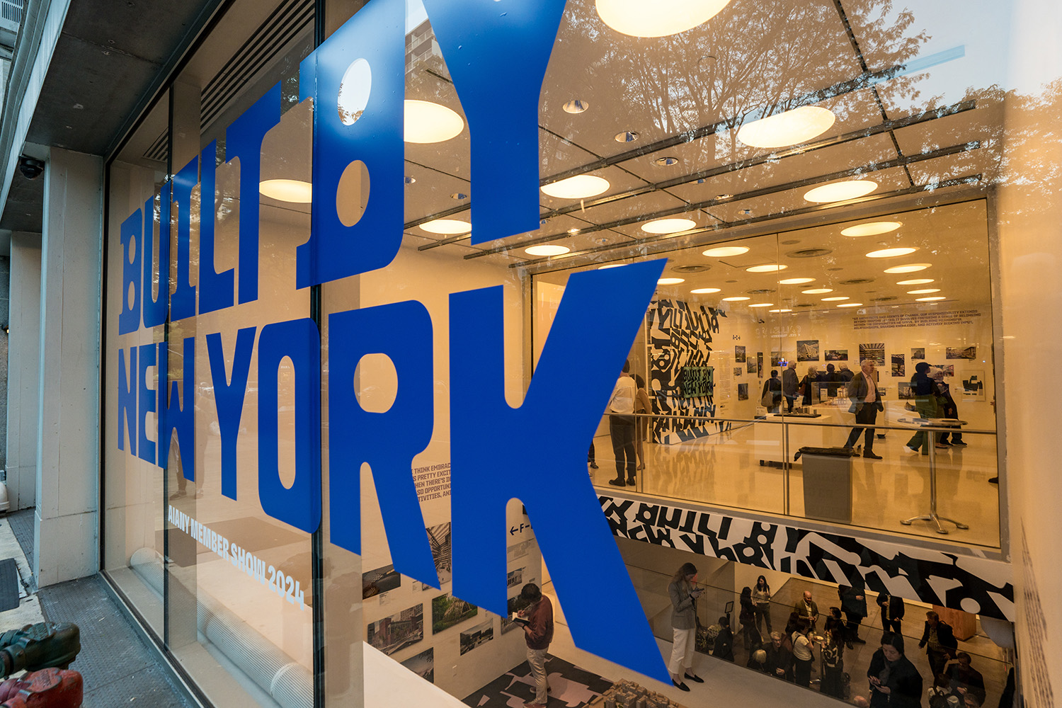 Bold blue text "Built by New York" on a glass window. Past it are three floors of exhibition gallery space.